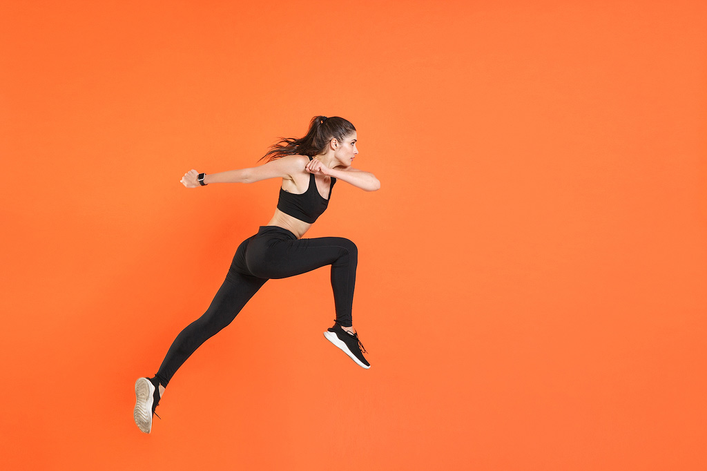 A woman poses as if she is running against a bright orange background.