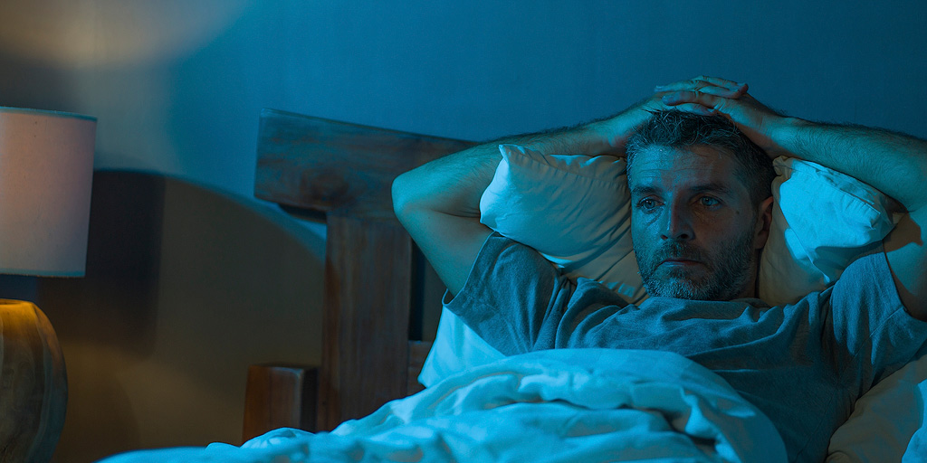 A man with a concerned facial expression experiences a sleepless night in a cinematically blue-lit bedroom.