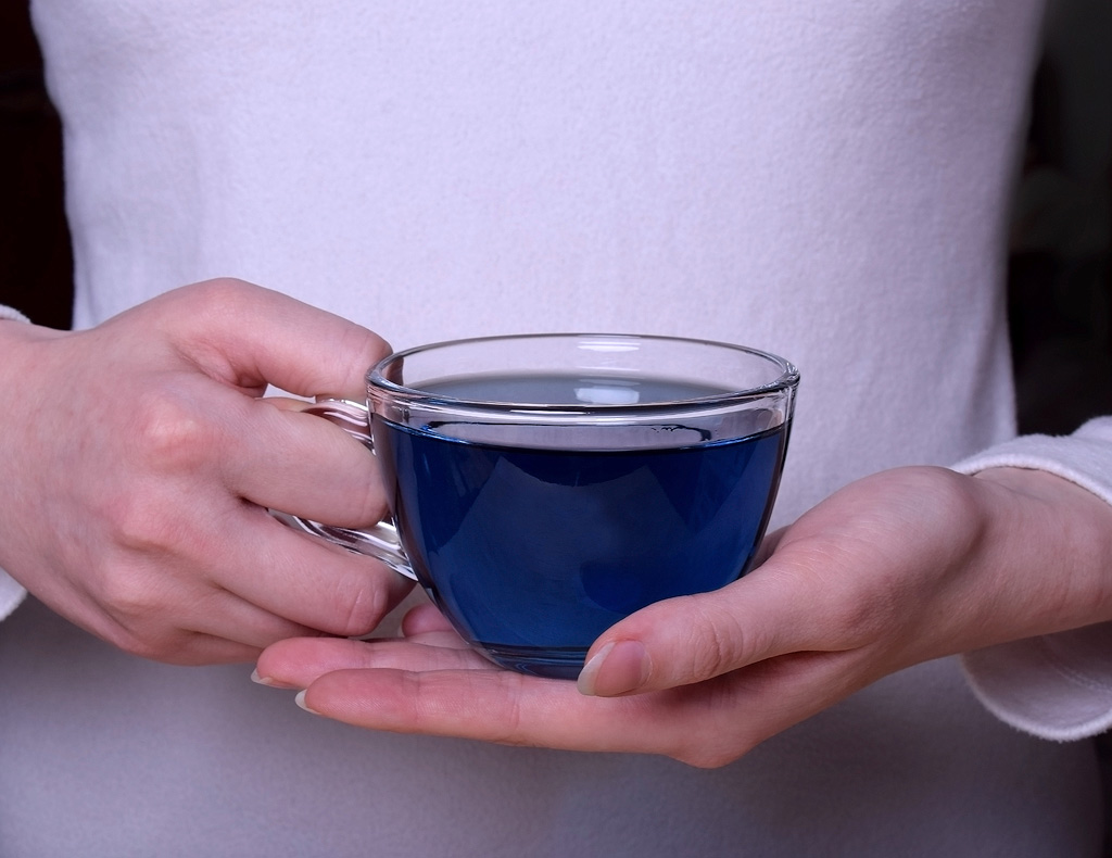 A woman holding a cup of blue tea in front of her stomach.