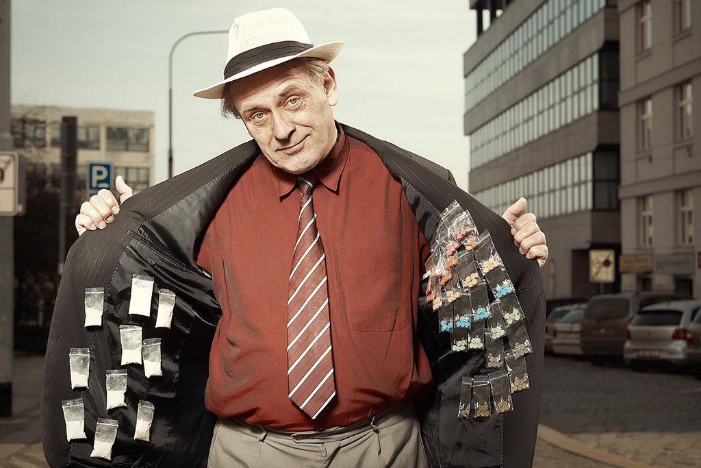 An older drug dealer with an open jacket displaying various drugs.